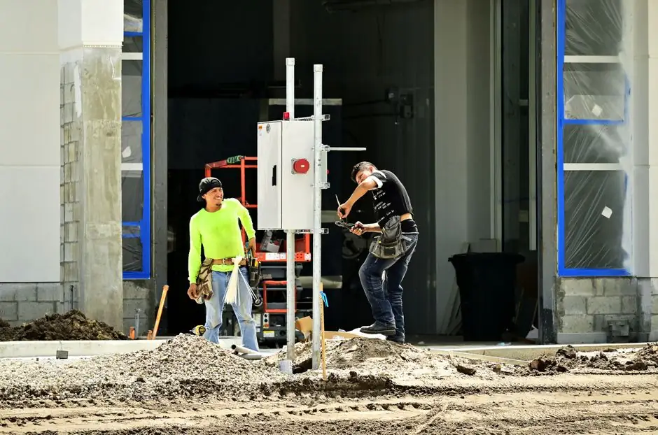 Two construction workers install an electrical panel outside a building site, wearing safety gear.
